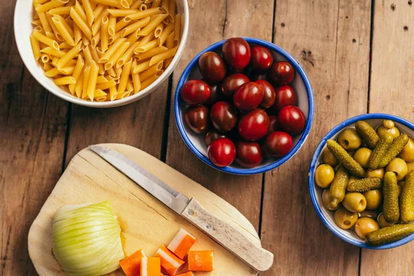 Eski ahşap bir masada makarna salatası için malzemeler, domates, zeytin, turşu, yengeç çubuğu, soğan ve makarna.