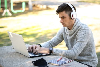 Genç adam parkta maskesiz kulaklıkla müzik dinlerken bilgisayarla çalışıyor.