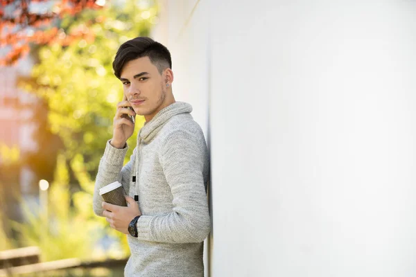 Parktaki gri kazaklı genç adam beyaz duvarın üstünde telefonla konuşuyor ve kahve içiyor. Kopyalama alanı