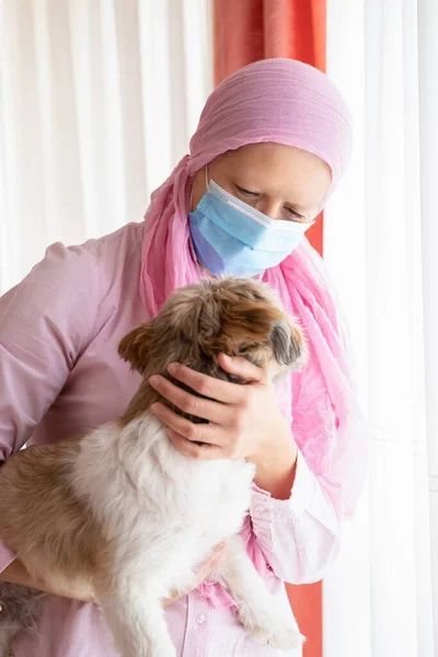 Kanserli kadın, pembe başörtüsü ve maskesi evcil hayvanına sarılıyor.