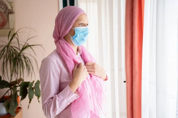 Kanserli kadın, pembe başörtüsü ve maskesi pencereden dışarı bakıyor.