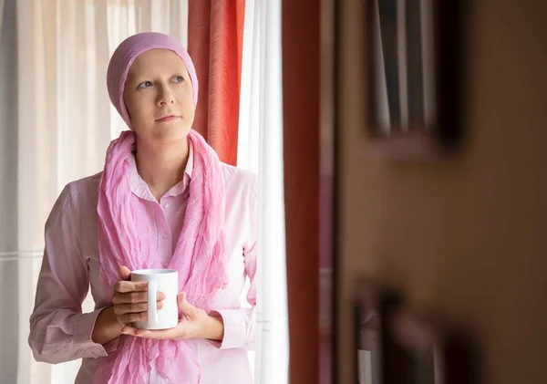 Kafasında pembe eşarp olan, kahve içip pencereden dışarı bakan kanserli bir kadın.