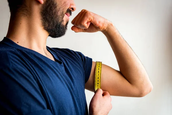 Bearded thin man measuring biceps, muscles of his left arm with a yellow tape measure. He\'s smiling, happy. Wearing blue t-shirt. White background.