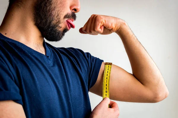 Bearded thin man measuring biceps, muscles of his left arm with a yellow tape measure. He's making a face; stretching tongue. Wearing blue t-shirt. White background.