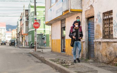 Bogota, Kolombiya 'da Corona virüsünün yayılmasını önlemek için maskeler takarak sokaklarda yürüyen Latin baba ve oğul. Yetişkin, çocuğunu omuzlarında taşır. Banliyöde güneşli bir gün. Fakir mahalle. 