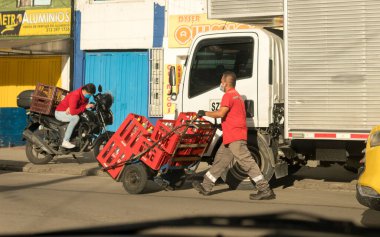Latin erkek kurye, Kolombiya, Bogota 'da Rojas Caddesi' nde bir el arabasında plastik bira şişesi sepetleri taşıyor. Corona virüsünün yayılmasını önlemek için maske takıyor. Yanında maske olan bir motosikletli..
