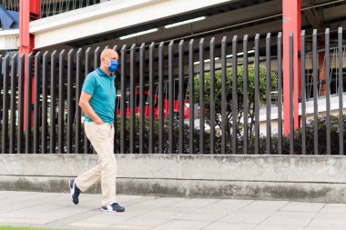 Bogota 'da sokakta yürürken koronavirüsün yayılmasını önlemek için maske takan orta yaşlı Latin bir adam. Kolombiya 'da. Arka taraftaki otoparkta. Mavi polo gömlek ve pantolon. 