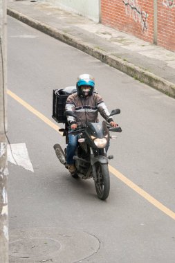 Kolombiya, Bogota sokaklarında motosikletini sürerken maske takan motosikletçi. Koronavirüs kapmaktan ve yayılmaktan kaçının. Siyah bisiklet, mavi kask. Kurye.