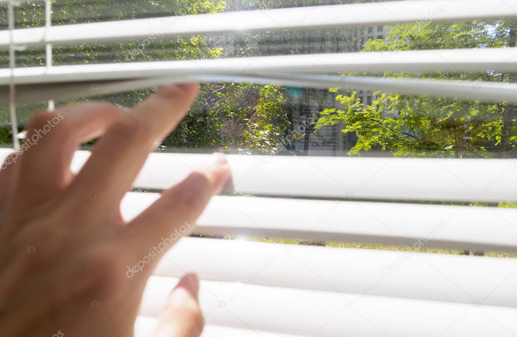 hand opens with fingers blinds, outside window there is sunlight and green trees, selective focus