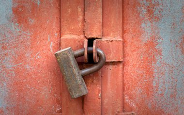 Kırmızı metal bir kapıya eski, paslı bir asma kilit takılmış. Yakın plan.