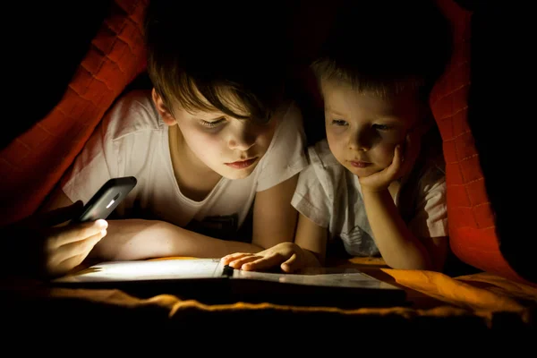 Children reading book with flashlight in tent.