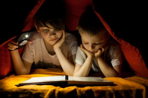 Children reading book with flashlight in tent.