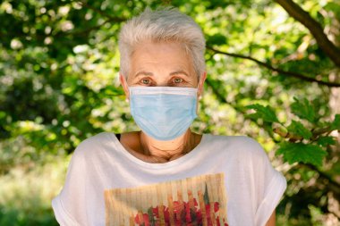 Yaşlı kadın tıbbi maskeyle doğanın tadını çıkarıyor. Kameraya bakıyorum..