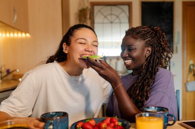 İki kadın sağlıklı bir kahvaltıdan zevk alıyor, biri diğerini beslerken diğeri de bir parça avokado tostunu paylaşıyor.