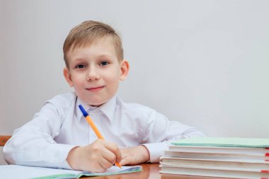 Çocuk masada oturuyor, elinde bir kalem, defterlerin ve ders kitaplarının yanında.
