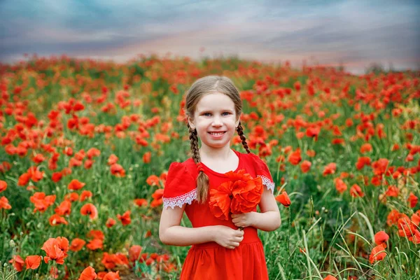 Dívka evropského vzhledu se dvěma náušnicemi v červených šatech stojí v krásném makovém poli proti — Stock fotografie