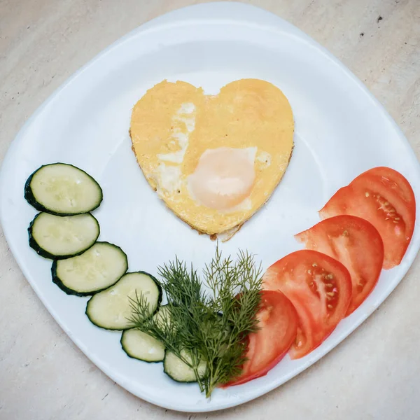 fried eggs in the form of a heart lie on a white plate, next to chopped dill, tomatoes and cucumbers
