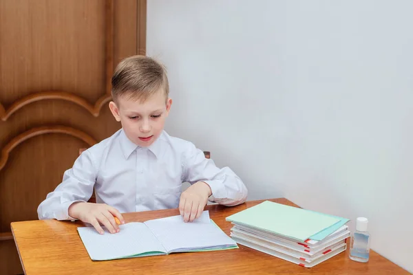 Ένα αγόρι της Ευρωπαϊκής εμφάνιση σε ένα λευκό πουκάμισο κάθεται σε ένα τραπέζι και κοιτάζει ένα σημειωματάριο — Φωτογραφία Αρχείου