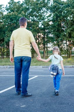 Baba ve kızı sokakta hafif giysiler içinde yürüyorlar, kız sırt çantası takıyor.