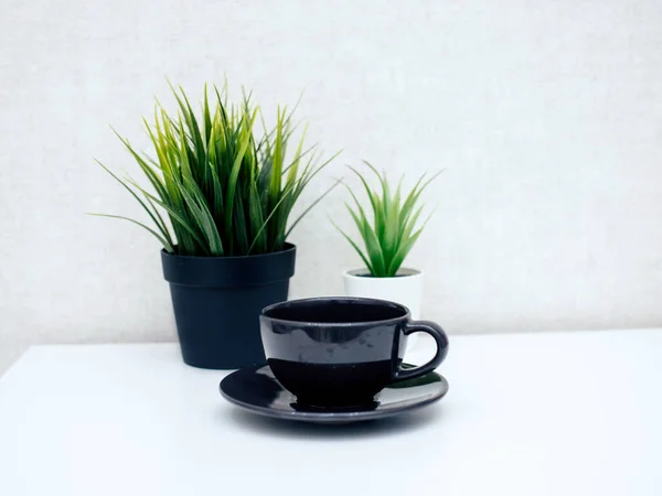 Sobre una mesa de luz hay una taza negra con una taza, y en el fondo dos flores en una olla — Foto de Stock