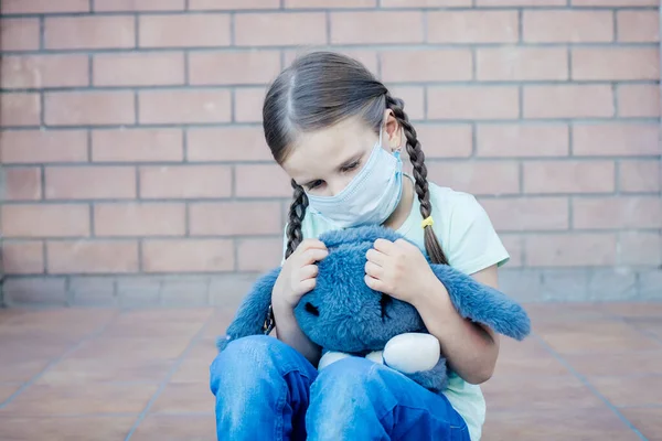 En el porche de la entrada se sienta una niña en una máscara con dos coletas y ojos tristes, sosteniendo una mochila en sus manos — Foto de Stock