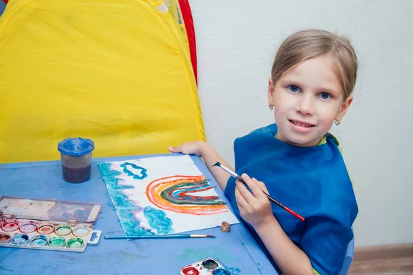 A menina está sentada à mesa e desenha com tintas coloridas . — Fotografia de Stock