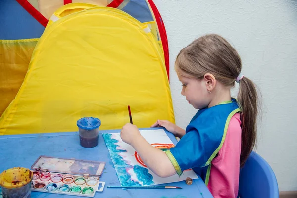 Το κορίτσι κάθεται στο τραπέζι και ζωγραφίζει με χρωματιστές μπογιές. — Φωτογραφία Αρχείου