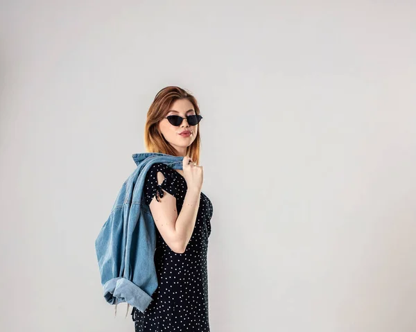 Una chica hermosa y suave con el pelo oscuro en un vestido negro con gafas y una chaqueta de mezclilla . — Foto de Stock