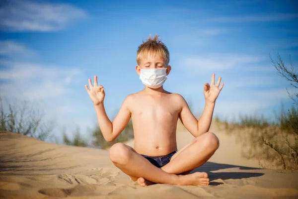 青い空と緑の茂みに対してマスクの砂の上に目を閉じたブロンドの少年が座っている. — ストック写真
