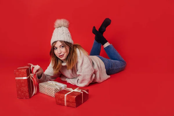 Dziewczyna w jasnym swetrze leży obok prezentów na czerwonym tle. — Zdjęcie stockowe