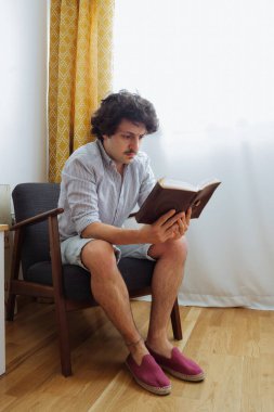 Okuma koltuğunda oturmuş kitap okuyan yakışıklı bir adam. Odaklanmış bir tavırla.