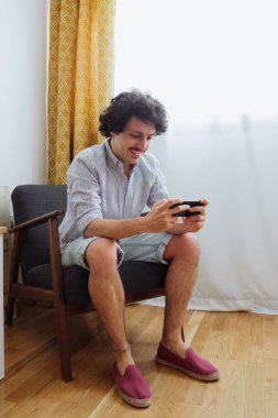 Elinde cep telefonuyla koltukta oturan yakışıklı genç adam rahat bir şekilde gülümseyip video oyunları oynuyor.