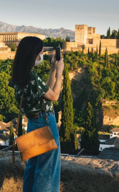 Maskeli genç bir kadın yeni normallik döneminde cep telefonuyla şehrin koronavirüsünü çekiyor. Seçici odaklanma. Kentsel Arapça arkaplan