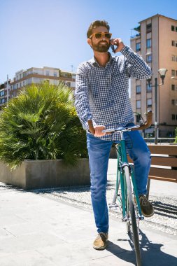 Güneş gözlüklü yakışıklı adam gülümsüyor ve cep telefonuyla konuşuyor. Tamir edilmiş vites bisikleti. Açık hava fotoğrafçılığı.