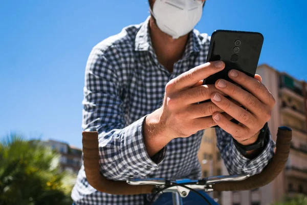 Hands of young man with mask with mobile phone and fixed gear bicycle in the street in time of coronavirus. Selective focus on hands. Hipster style.