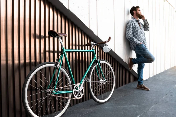Attractive man smiling and standing on a two-color modern wall next to his bike while calling with his cell phone. Horizontal photography, hipster style.