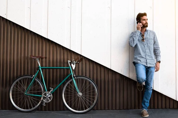 Attractive man standing on a two-color modern wall next to his bike while calling with his cell phone. Horizontal photography, hipster style.