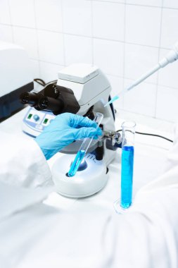 Bilim adamı, pipetle mavi örneği tıbbi mikroskop altında görmek için test tüpüne dönüştürüyor. Virüs salgını.