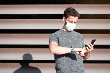 Tıbbi maskeli yakışıklı adam koşmaya başlamadan önce cep telefonuna bakıyor. Açık alanda modern arkaplan.