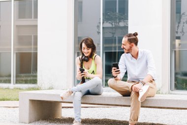 Çift bankta oturuyor gülümsüyor ve cep telefonlarına bakıyor..