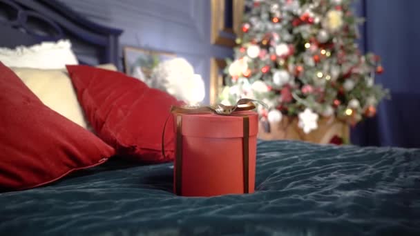Geschenk in einer roten Schachtel mit Schleife auf dem Bett liegend. Ferien. Geschenk für das neue Jahr auf dem Hintergrund des Weihnachtsbaums — Stockvideo