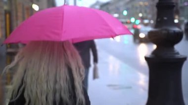 Şehir merkezinde yağmurda pembe bir şemsiye altında yürüyen bir kız. Şemsiyenin altında yürüyen sarışın kız.