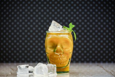 Kafatası şeklindeki bir bardaktaki şeftali suyu mermer bir tahtada, arka planda da buz parçaları var. Taze şeftali suyu, buz ve kafa şeklinde olağandışı bir kupa. Cadılar Bayramı zombi kokteyli