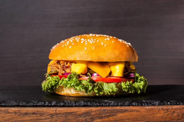 用猪排做的汉堡躺在一块石头做的木板上.还有泡菜，奶酪，红洋葱，芥末和新鲜沙拉叶。后面的黑色木制背景 图库图片