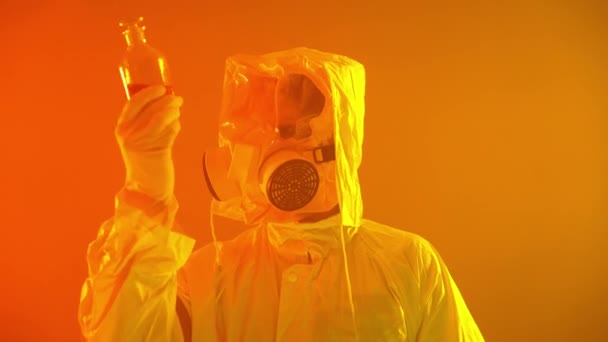 Man in een geel beschermingspak met rode vloeistof in de erlenmeyer. Man met gasmasker Uitvinding van het vaccin. Ecologie en biogevaarprobleem. Gele achtergrond met zware rook. COVID-19, coronavirus. — Stockvideo