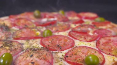 Macro shot of freshly baked pizza topped with sliced tomatoes, green olives, melted cheese and herbs, with visible steam rising. High quality 4k footage