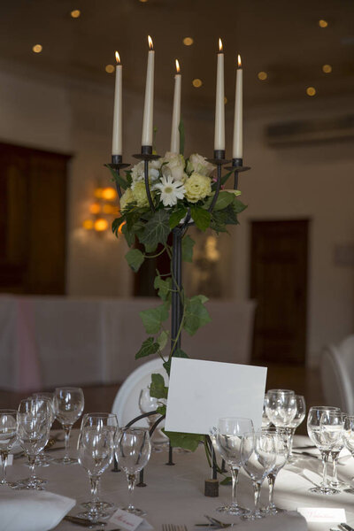 elegantly served with glasses, cutlery and decorated with flowers tables with white tablecloths and burning candles in candlesticks in the blurred background of the fashionable restaurant hall.