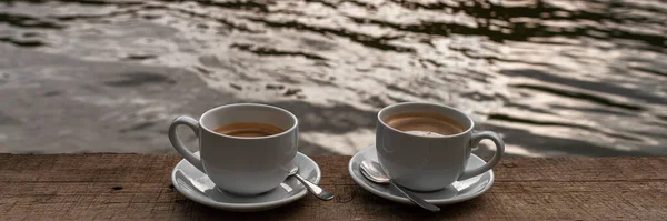 Baner dwie filiżanki kawy na drewnianym stole z bocznym jeziorem lub rzeką — Zdjęcie stockowe