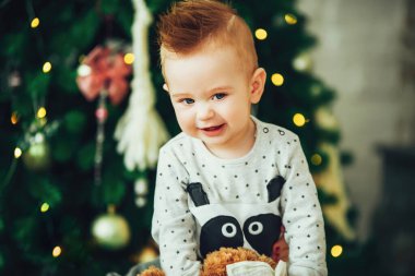 Bebek 1 yaşında, üzerinde Noel ağacı olan pijama giyiyor ve arka planda ışıklar var.