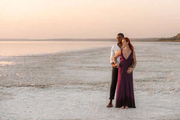 Portrait of adorable young man hugging his girlfriend from behind while standing together. Happy mixed-race millennial couple enjoying a day at beach.